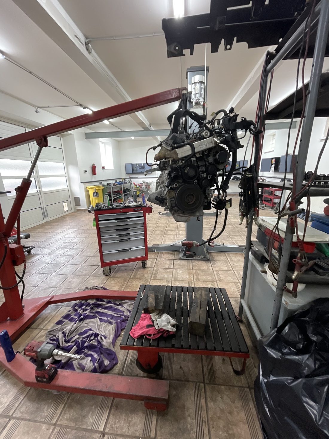 Dílna se světlou dlažbou, uprostřed motor zavěšený na červeném dílenském jeřábu, vzadu vozík s nářadím
